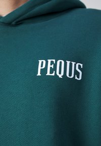 Žalia džemperis su minkšta tekstūra ir spausdintu baltu logotipu, kuris per krūtinę skaito "PEQUS". Paprastas, kasdienis dizainas.