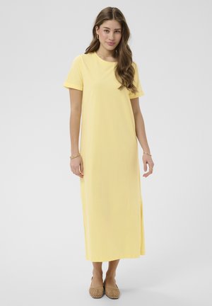Junge Frau mit langen braunen Haaren, die ein schlichtes gelbes Maxikleid mit kurzen Ärmeln und beigen geflochtenen flachen Schuhen trägt, steht vor einem hellen Hintergrund.