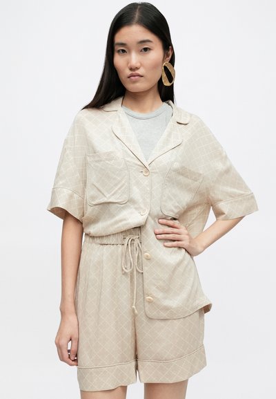 Béžová patterned košeľa s krátkym rukávom a dvoma prednými vreckami, skombinovaná s elastickými šortkami na šnúrku, ktoré sú farebne zladené. Hladká tkanina, uvoľnený strih a jemný geometrický vzor.