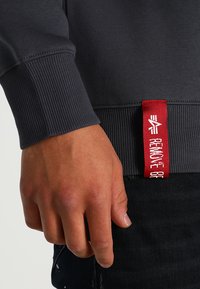 Sötétszürke pulóver bordás mandzsettával és aljjal. Piros címke fehér szöveggel. A kéz a szegély mellett helyezkedik el.