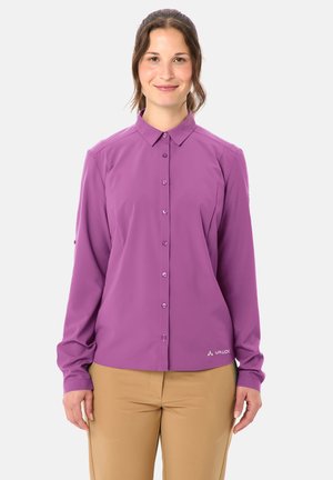 Vrouw staand en glimlachend, draagt een lange mouwen violet blouse met knopen en beige broek tegen een effen lichte achtergrond.