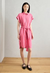 Abito camicia di lino rosa con maniche corte, chiusura a bottoni frontale, cinturino in vita e spacchi laterali. Indossato con scarpe nere basse su un pavimento in legno.