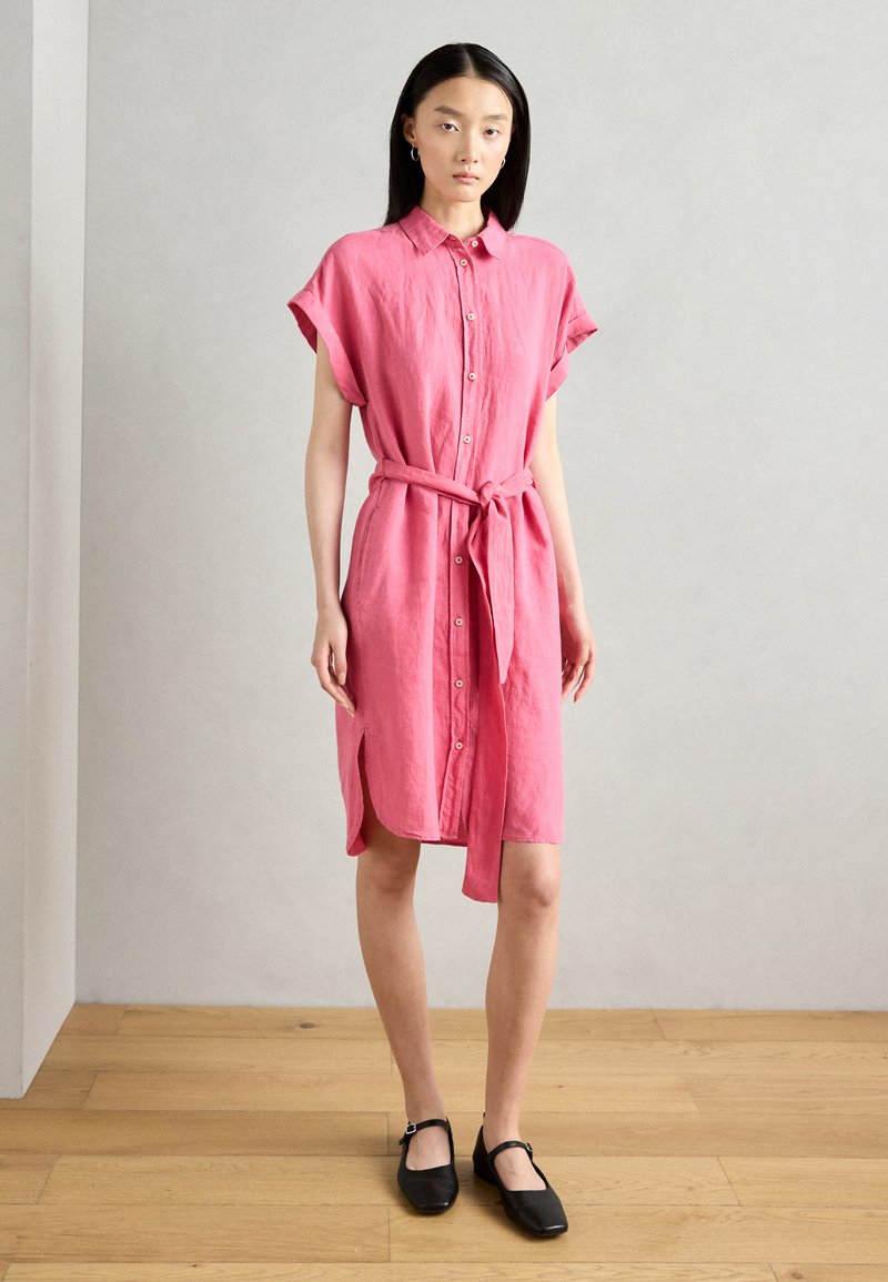 Abito camicia di lino rosa con maniche corte, chiusura a bottoni frontale, cinturino in vita e spacchi laterali. Indossato con scarpe nere basse su un pavimento in legno.