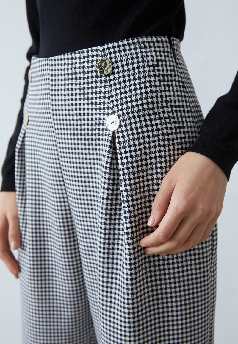 Pantaloni a vita alta in un motivo a scacchi bianco e nero, caratterizzati da pince e bottoni lucidi in oro e bianco. Tessuto morbido con una vestibilità strutturata.