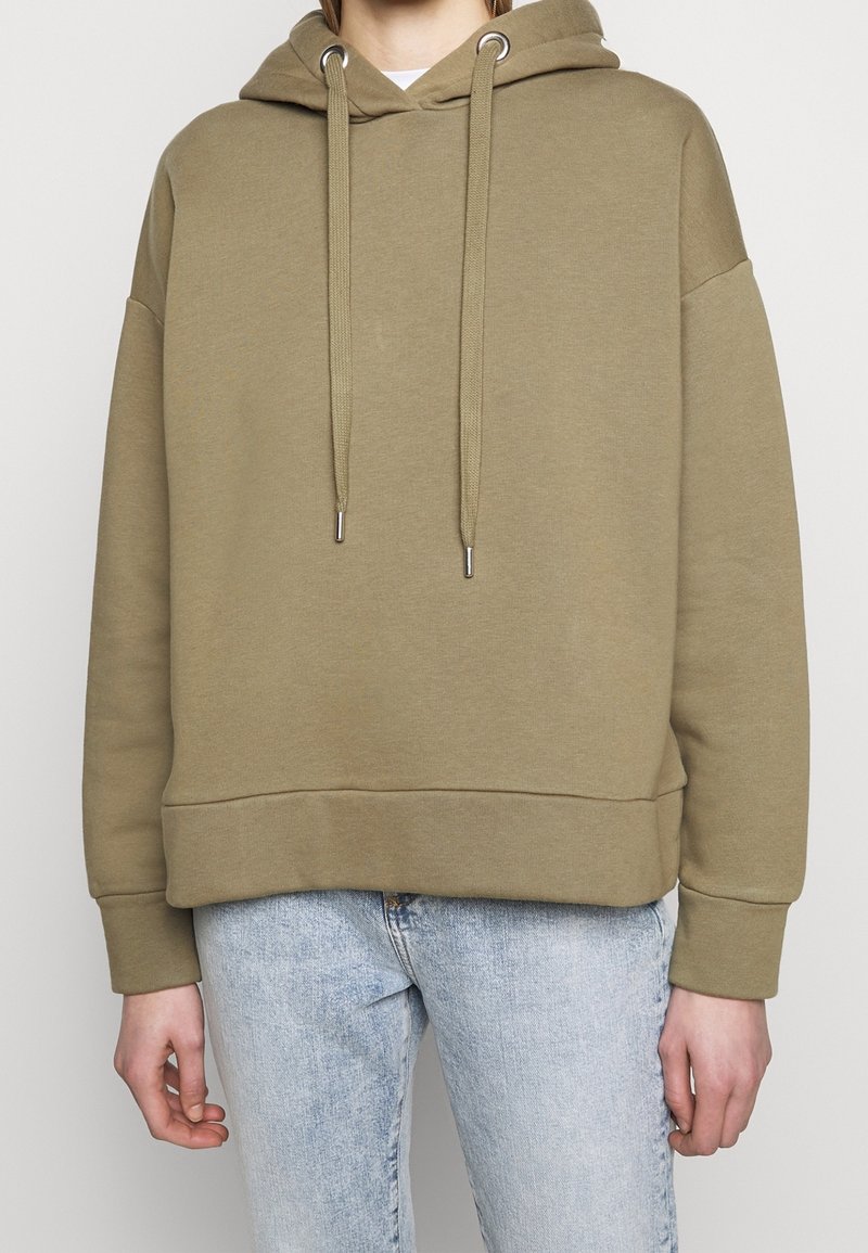 Personne portant un sweat à capuche vert olive uni avec cordons et un jean bleu clair, montré du cou aux hanches sur un fond neutre.