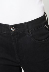 Jeans de mezclilla negros con un botón brillante y decorado en la cintura, con pasadores para el cinturón estándar y bolsillos frontales. Textura suave, diseño ajustado.