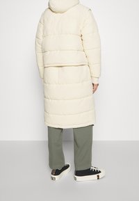 Persona vestida con un abrigo de plumas largo color crema, pantalones de pierna ancha verde oliva y zapatillas bajas negras con suelas blancas, de pie de espaldas.