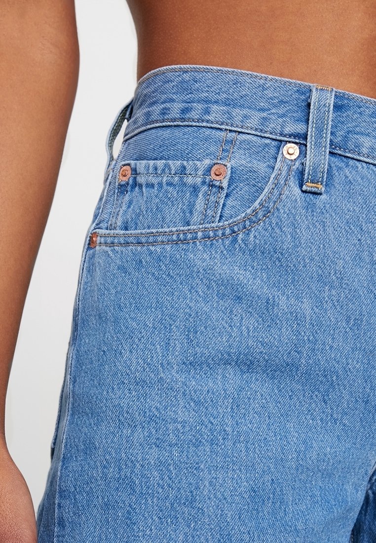 Närbild på en persons midja i högmidjade ljusblå denimjeans med synlig framficka och kopparnitar.