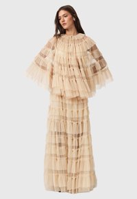 Lagerad beige tyllklänning med volanglager, långa ärmar och rundad halsringning. Den genomskinliga texturen förstärker det flytande, lätta utseendet.
