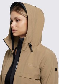 Giacca impermeabile beige con cappuccio, dettagli neri, zip frontale e logo sul petto. Presenta un tessuto testurizzato e cordino regolabile.