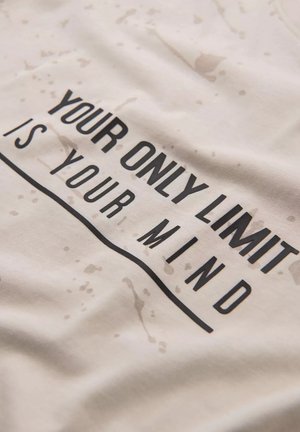 Valkoinen puuvillapaita, jossa on musta teksti "AINOA RAJA ON MIELESI", kevyillä roiskuviivoilla ja sileällä pinnalla.