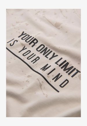 Valkoinen puuvillapaita, jossa on musta teksti "AINOA RAJA ON MIELESI", kevyillä roiskuviivoilla ja sileällä pinnalla.