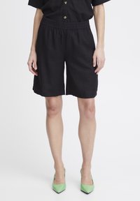Shorts en lin noirs avec une taille élastique, dotés de poches latérales et d'une coupe décontractée. Les chaussures sont vertes et ont un bout pointu.