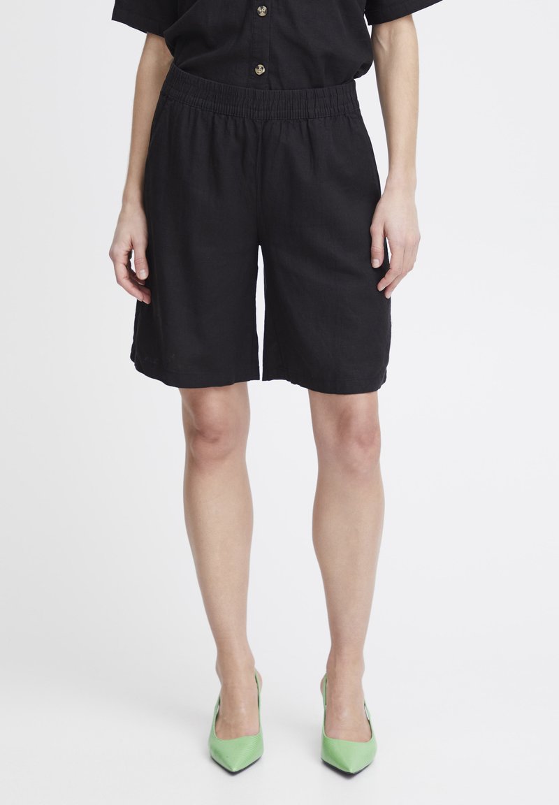 Shorts en lin noirs avec une taille élastique, dotés de poches latérales et d'une coupe décontractée. Les chaussures sont vertes et ont un bout pointu.