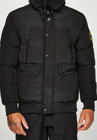 Sort dunjakke med lynlås og knaplukning, to forlommer, ribbede manchetter og lagdelt struktur. Har en hætte og et logopatch.