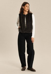 Vrouw met lang bruin haar in een zwart mouwloos bontvest over een wit longsleeve shirt, zwarte broek en bruine instappers, staand met haar hand in de zak.