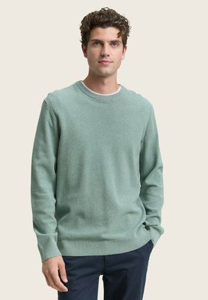 Suéter de punto verde claro con un cuello redondo y puños acanalados. La textura parece suave, con un sutil patrón horizontal. Llevado con pantalones azul marino.