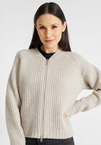 Beige gebreide cardigan met een ribbelstructuur, raglanmouwen en een voorrits. Heeft een hoge kraag en een aangesloten zoom.