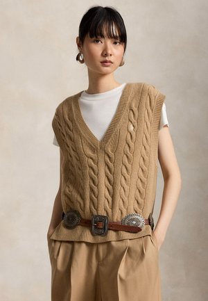 Kvinde iført en beige kabelstrikket vest over hvid T-shirt, lysebrune plisserede bukser og et brunt bælte med store sølvconchos, stående med hænderne i lommerne.