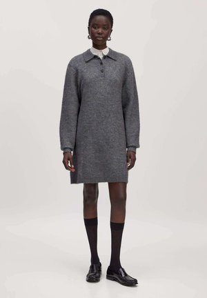 Frau in einem grauen, langärmligen Strickkleid, schwarzen knielangen Socken und schwarzen Loafern, steht vor einem schlichten hellen Hintergrund.