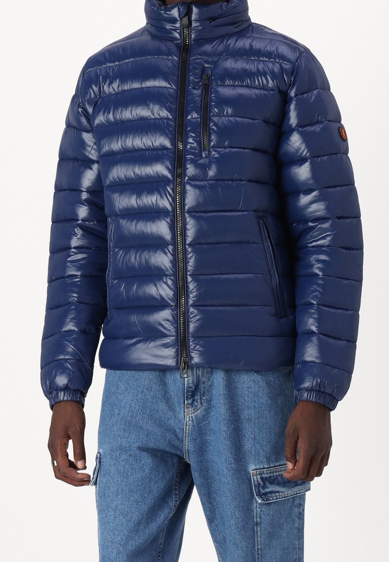 Marineblå pufferjakke med quiltet design, høj krave, frontlynlås og en brystlomme; lavet af blankt, letvægtsmateriale.