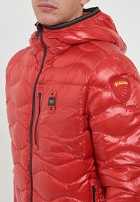 Giacca a vento rossa con una texture lucida e trapuntata, dotata di zip frontale, cappuccio e una toppa con logo sulla spalla. Accenti neri e gialli.