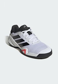 Bela športna copatka s črnimi poudarki in tremi črnimi črtami. Zgornji del iz teksturirane mreže, gumijasti podplat in rdeč detajl na prstih. Vključeni so vezalke.