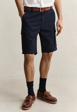 Uomo che indossa pantaloncini cargo blu navy, cintura di pelle marrone, calzini neri e scarpe casual di pelle marrone, in piedi su uno sfondo chiaro.