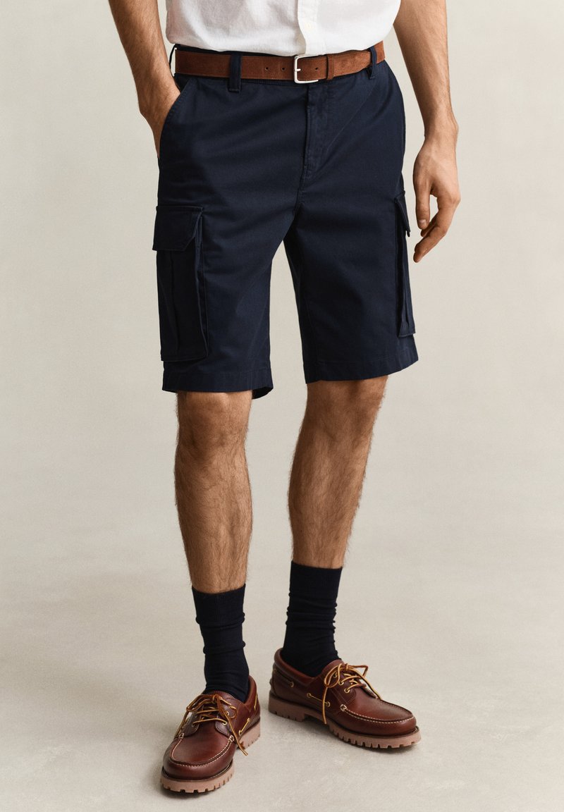 Uomo che indossa pantaloncini cargo blu navy, cintura di pelle marrone, calzini neri e scarpe casual di pelle marrone, in piedi su uno sfondo chiaro.