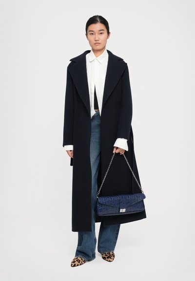 Donna che indossa un lungo cappotto scuro sopra una camicia bianca con bottoni e jeans blu, tiene una borsa blu con catena argentata e indossa scarpe con stampa leopardata.