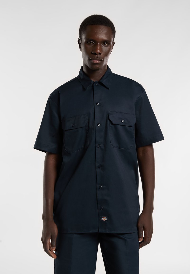 Camisa de manga corta, de color azul marino oscuro, con dos bolsillos en el frente, cuello abotonado y tela ligera. Presenta una etiqueta con el logo cerca del dobladillo.