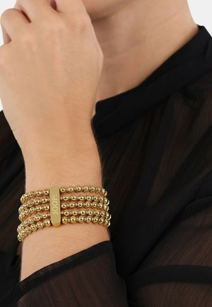 Bracciale con perline d'oro a più fili e una piastra centrale incisa con "JOOP!" La texture liscia e lucida completa un top nero trasparente.