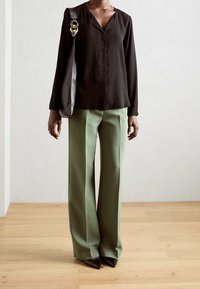 Camicia nera a maniche lunghe con scollo a V, abbinata a pantaloni verdi a vita alta e gamba larga, insieme a décolleté nere con punta. Accessori dorati sulla borsa.