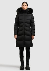 Mustast õhukindlast materjalist talvejope eemaldatava karvaga kapuutsiga, tikandiga, ees tõmblukk ja küljetaskud, kombineeritud mustade retuuside ja paeltega saapadega.