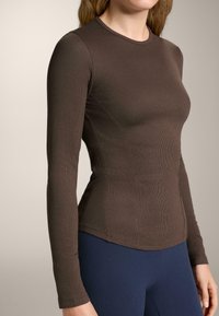 Smeđa majica dugih rukava u rebrastoj teksturi s uskim krojem. Ima okrugli izrez i glatke šavove, nosi se preko tamnoplave tajice.