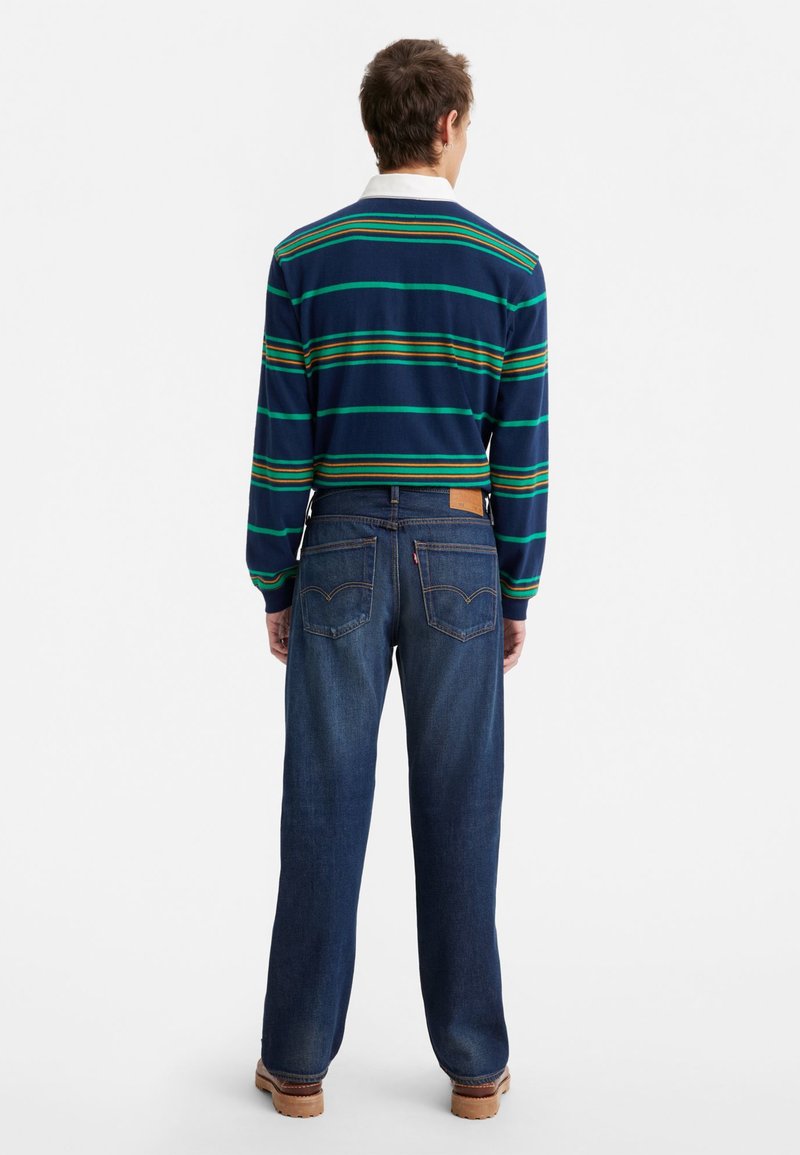 Blau gestreiftes Langarmhemd mit grünen und orangen Streifen, kombiniert mit dunkelblauen Jeans und braunen Schuhen. Das Hemd hat einen weißen Kragen.