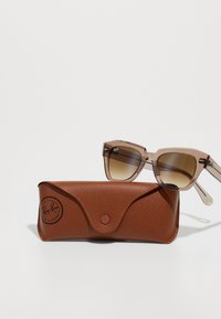 Transparente brune Ray-Ban solbriller med gradientlinser som hviler på et brunt lær Ray-Ban-etui mot en hvit bakgrunn.