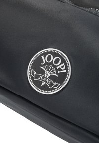 Borsa nera in nylon con un logo rotondo bianco con la scritta "JOOP! JEANS" e un design decorativo. Texture liscia con chiusura a zip.