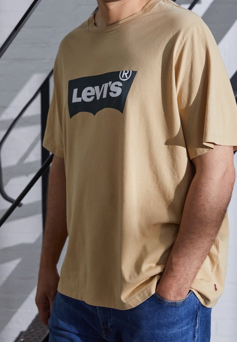 T-shirt en coton beige avec un logo noir Levi's, coupe décontractée, manches courtes et encolure arrondie, associé à un jean en denim bleu.