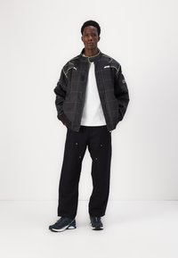 Black jacket with white grid pattern and green accents, white t-shirt, black pants with pockets, and navy athletic shoes.