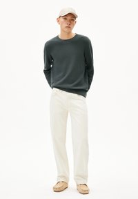 Pull en maille vert foncé, jean beige et chaussures slip-on marron clair. Le modèle porte une casquette beige clair au design simple.