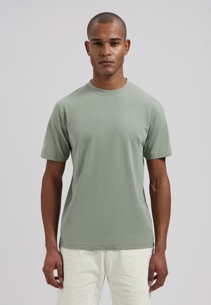 Man with short hair wearing a plain light green short-sleeve t-shirt and white pants, standing against a plain gray background.