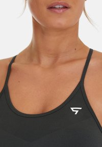 Schwarzes Sporttanktop mit dünnen Trägern, rundem Halsausschnitt und einem weißen Logo. Der Stoff hat eine glatte Textur mit einem leichten Glanz.