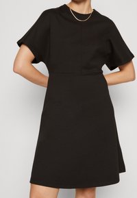 Czarna sukienka z krótkimi rękawami, okrągłym dekoltem, uszyta z miękkiej tkaniny, o luźnym kroju i długości do kolan, z minimalnie widocznymi szwami.