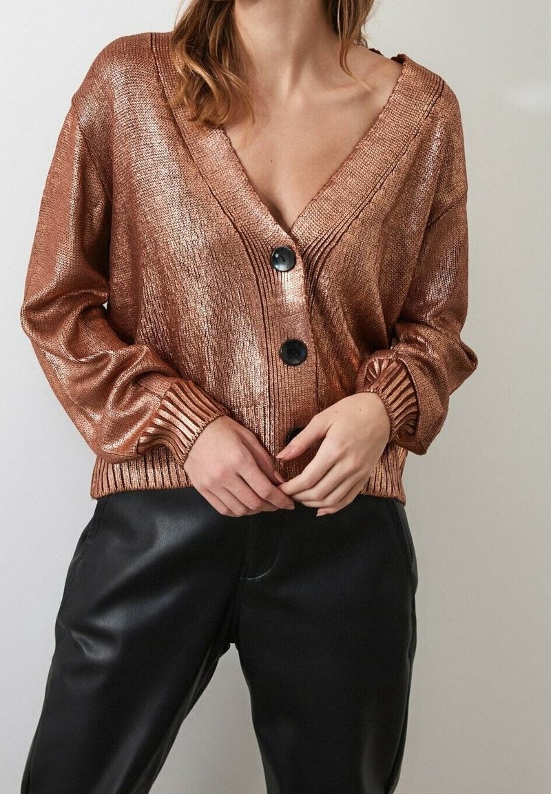 Cardigan de punto metálico en cobre con escote en V profundo, botones negros, puños acanalados y dobladillo inferior, combinado con pantalones de cuero negros.