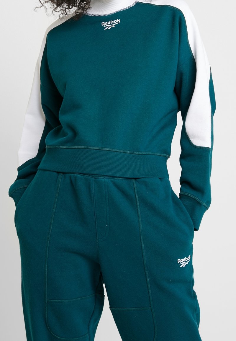 Personne portant un sweat-shirt Reebok bleu sarcelle et blanc assorti avec un pantalon de survêtement, les mains dans les poches, faisant face à l'avant.