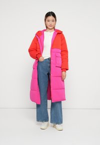 Cappotto lungo imbottito con blocchi di colore rosso e rosa, bottoni a scatto e tasche laterali, abbinato a jeans ampi blu e scarpe da ginnastica bianche.