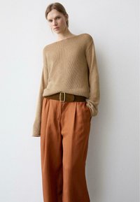 Kvinde iført en løs beige strikket trøje, stukket ned i højtaljede, rustfarvede bukser med vide ben og et brunt bælte.