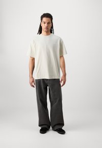 Beige t-shirt med kort ärm och en subtil logga, kombinerad med löst sittande mörkgrå jeans och svarta skor. Enkel och avslappnad design.
