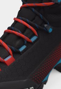 Közeli kép egy fekete sportcipőről, piros és fekete fűzőkkel, kék varrással és piros hálós részletekkel az oldalán.